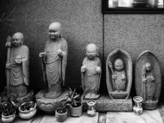 Monochrome image of five distinct Buddhist statues on a shelf with offerings.