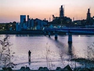 Twilight Solitude Solitary figure fishing by a serene inlet against a twilight industrial cityscape.
