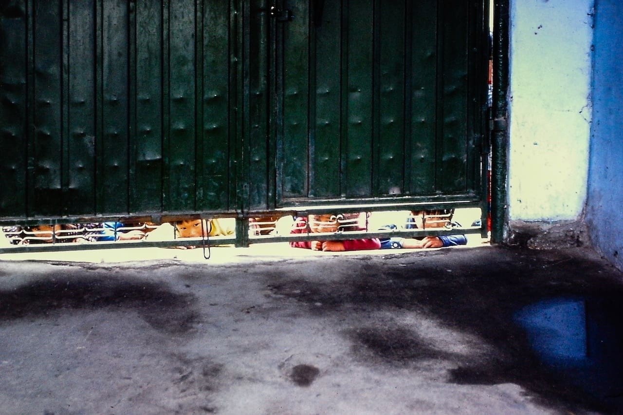Curious Korean children Peering from Weathered Green Urban Gate Curious Korean children peeking from beneath an aged green metal gate in a vibrant urban setting.