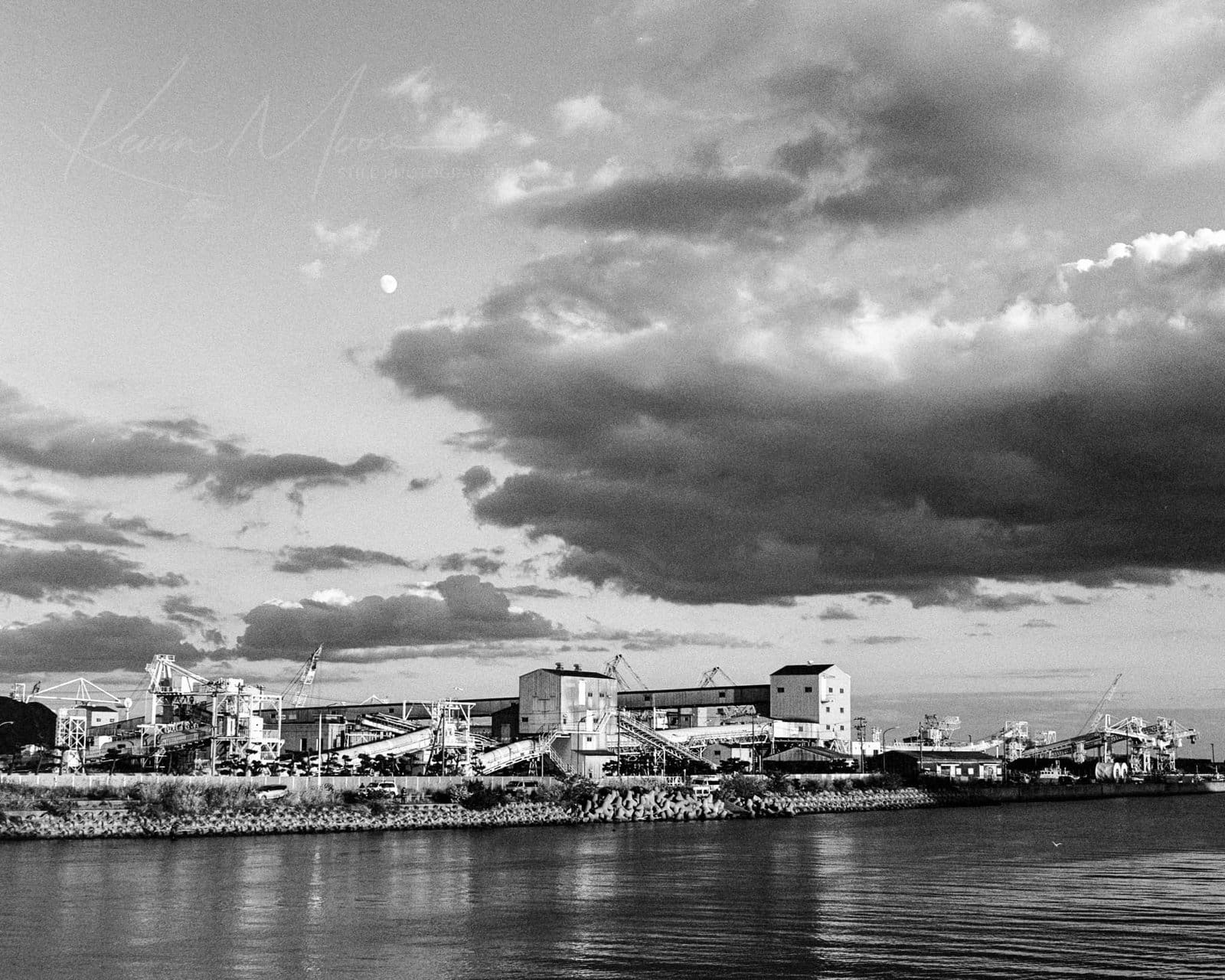 Industrial Riverside at Dusk: Factories and Moonlit Sky Industrial riverfront with factories, cranes, and reflective water under a cloudy sky with distant moon.