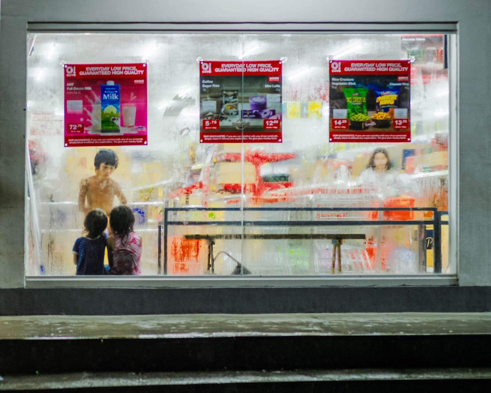 Rainy window art: two children at a fogged grocery store window at night in the Philippines, red posters blurred by rain